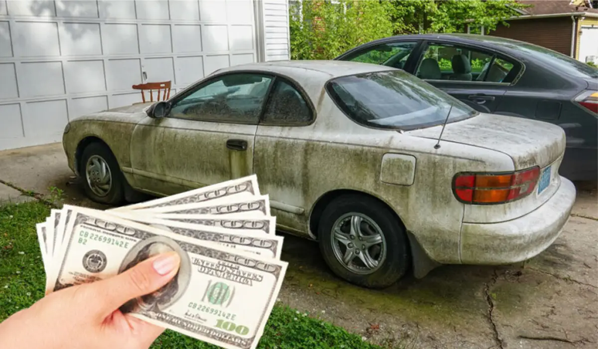 A mans hand holiding a five hundred dollar bills in front of the junk car.