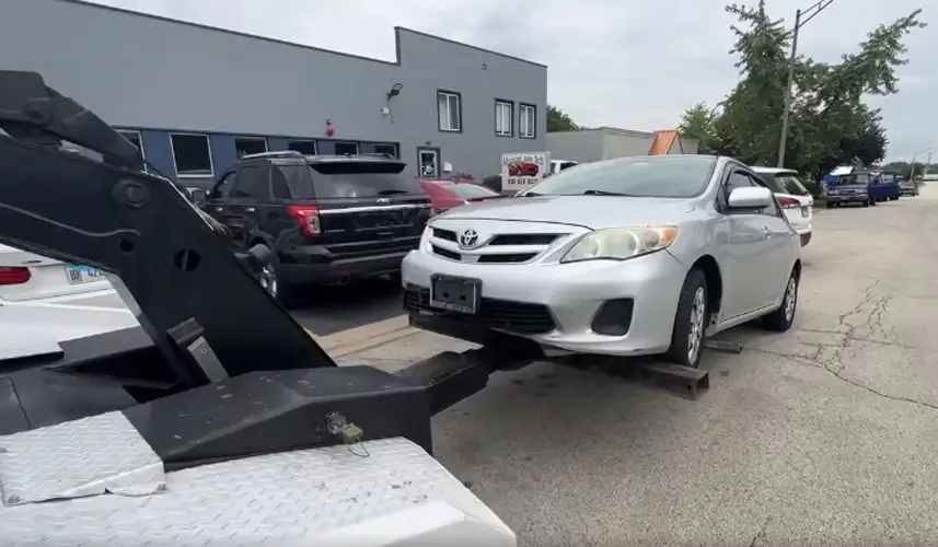 Car removal service towing a white Toyota sedan using a professional tow truck on an urban street.