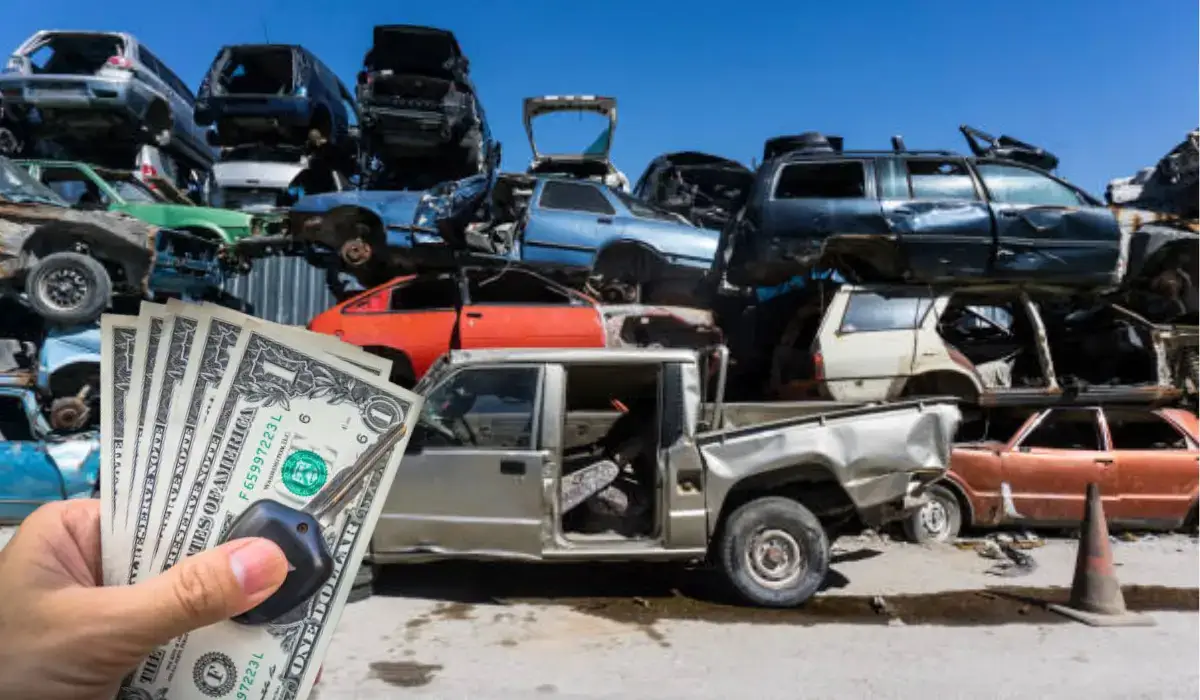 A hand holding cash and a car key in front of a junkyard filled with damaged vehicles.