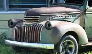 A heavily rusted, abandoned junk vehicle with missing headlights and faded paint, sitting in overgrown grass.