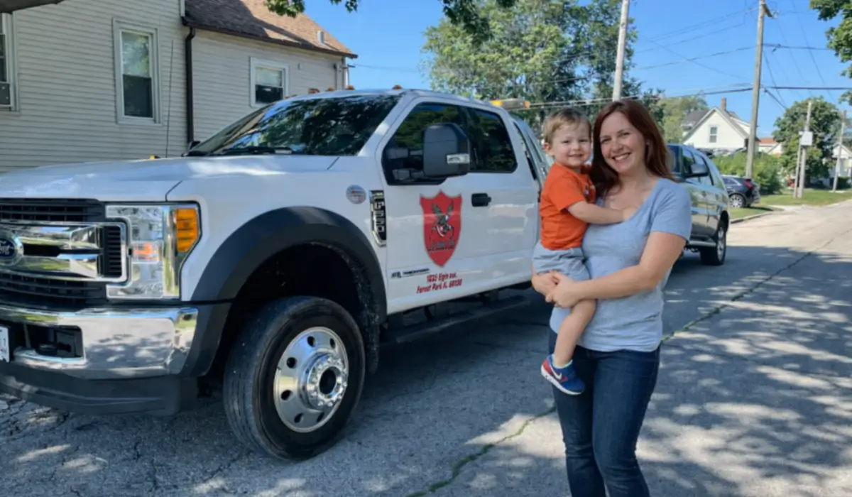 Single mother holding child beside tow truck during junk car removal pickup in Willowbrook neighborhood driveway