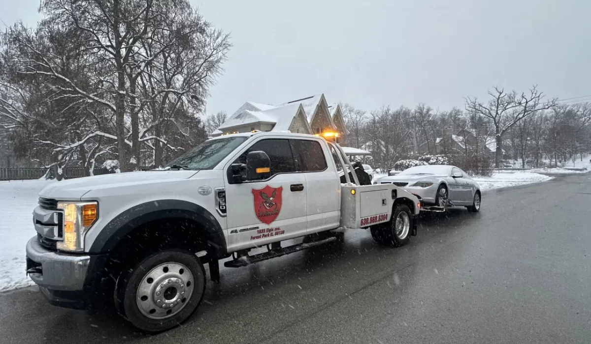 Tow truck hauling a junk car in winter during snowfall on a residential Bolingbrook street.